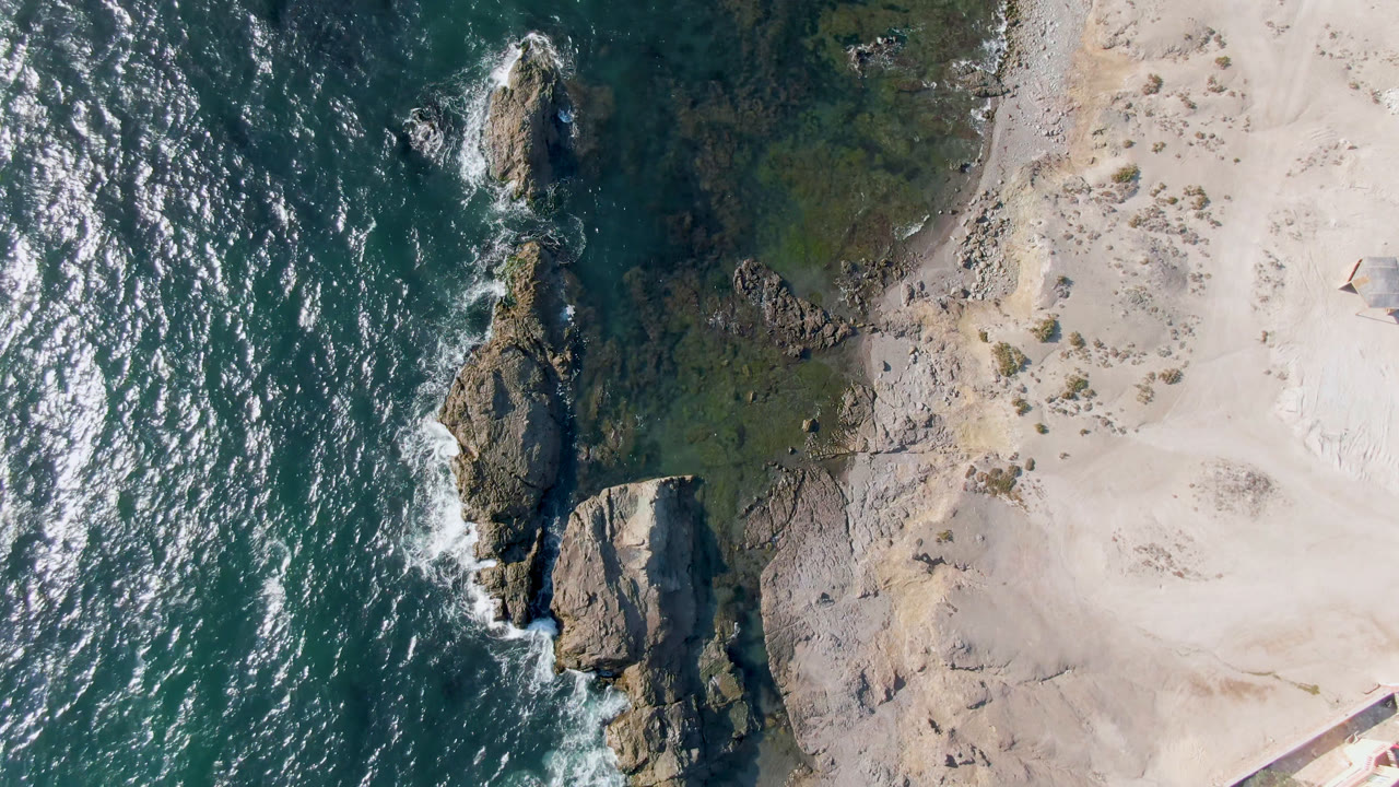 Aerial view over Bahia Ascuncion showing teal ocean on the left, pale sand on the right, jagged rocks at the shore, and no visible people.