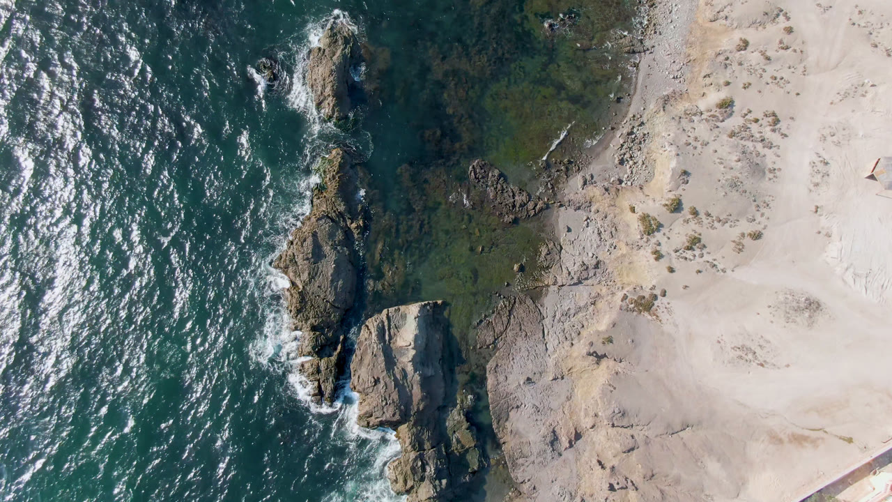 Overhead frame from Bahia Ascuncion with white surf wrapping around a vertical line of rocks beside a wide pale beach, with no visible people.