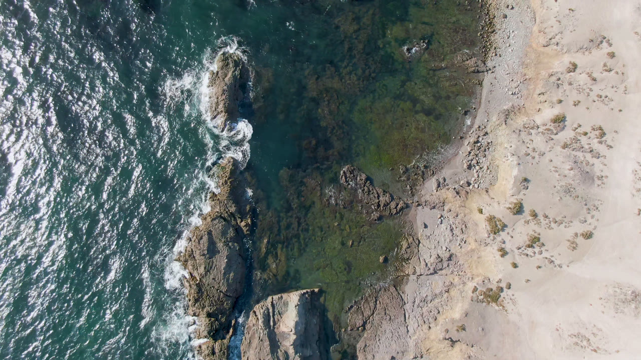 Overhead view of Bahia Ascuncion shoreline with teal ocean on the left, exposed rocks near center, pale sand on the right, and no visible people.