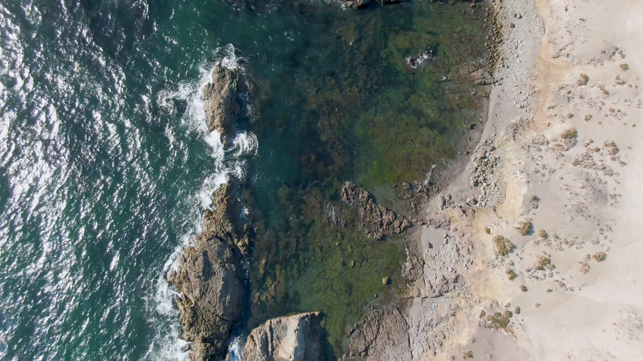 Aerial frame over Bahia Ascuncion shows white foam wrapping the central rock line, darker teal water to the left, shallow green water near shore, and no visible people.