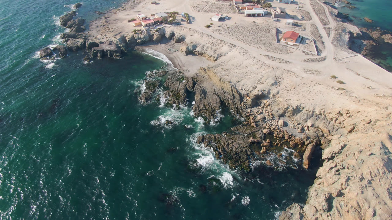 Drone view over Bahia Ascuncion, Baja California Sur, with dark water and white surf hitting rocks below pale sand and small buildings, no visible people.