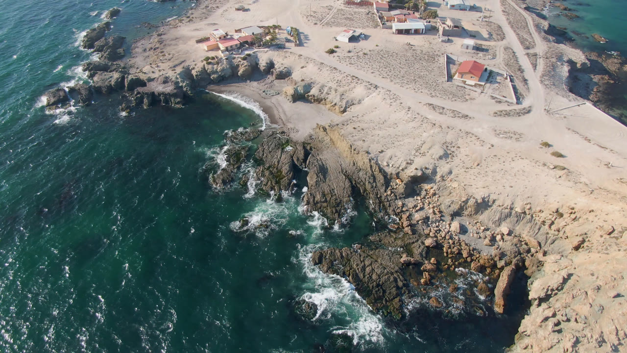 A high aerial frame in Bahia Ascuncion shows surf curling around a rocky cove, pale beach tracks, and red-roof coastal buildings, no visible people.