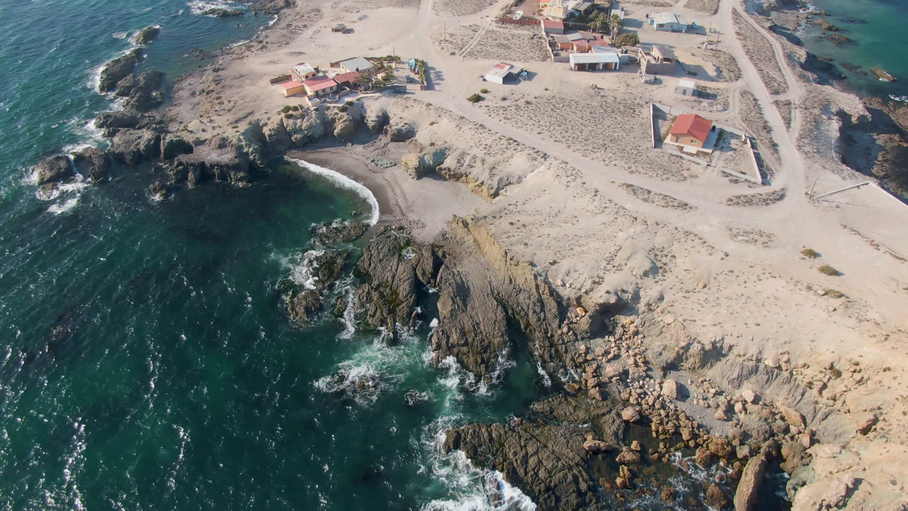 Drone view of a sandy coastal point with rocks, surf, road tracks, and scattered buildings.