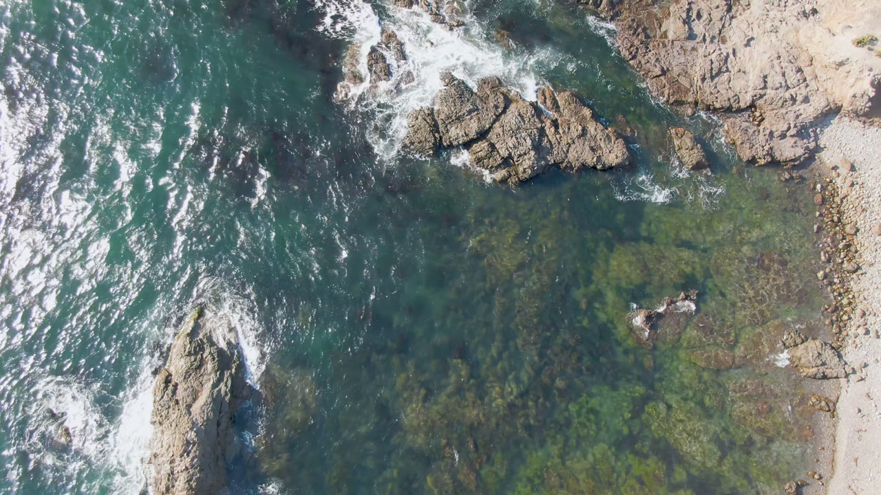 Overhead frame of Bahia Ascuncion shoreline with tan rocks, clear blue-green water, white surf foam, and no visible people.