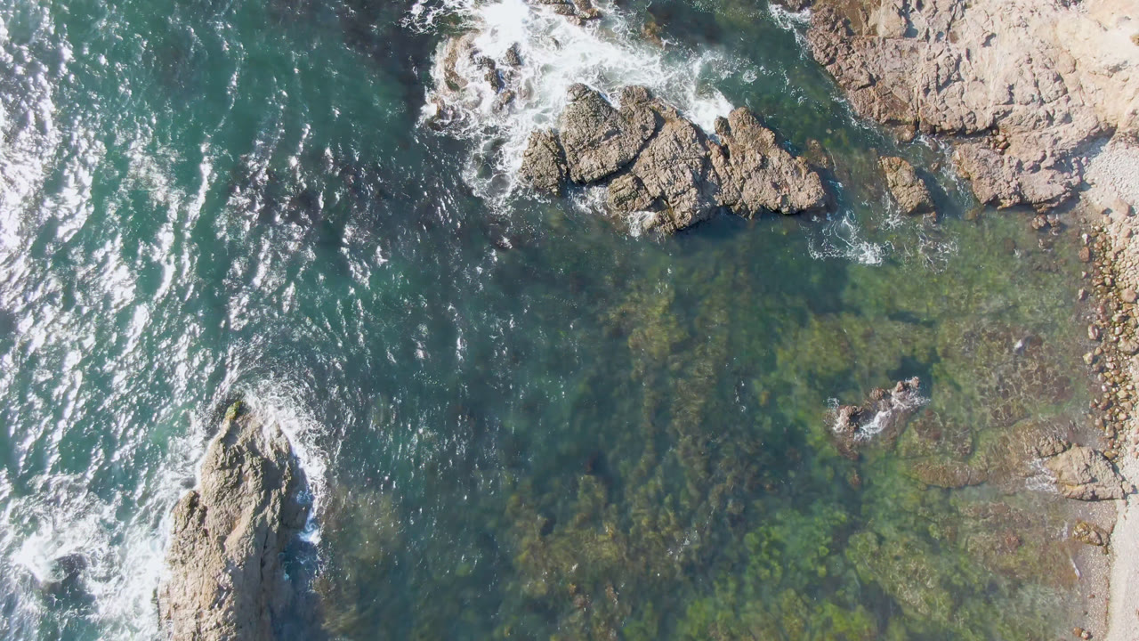 Drone frame over Bahia Ascuncion with white surf wrapping around a large tan rock, transparent shallows, darker underwater patches, and no visible people.