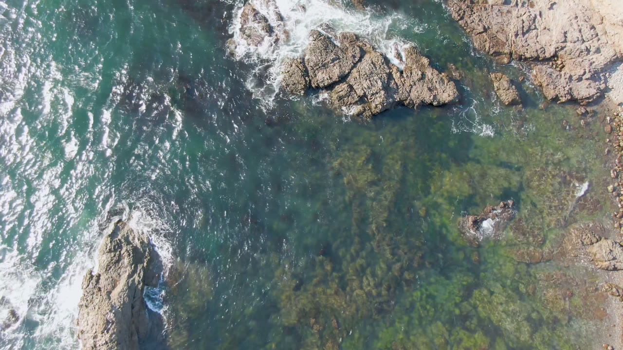 Top-down coastal water with rocks, foam, and submerged dark patches