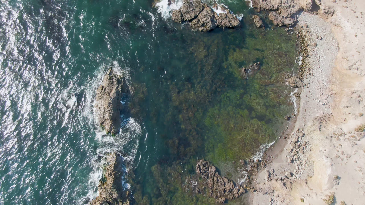 Overhead view of Bahia Ascuncion shoreline with teal water on the left, pale sand on the right, rock formations in the surf, bright water reflections, and no visible people.