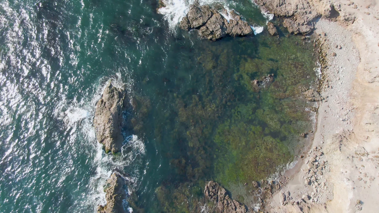 Aerial frame over Bahia Ascuncion with surf curling around central rocks, green shallows close to the beach, pale sand at the right edge, and no visible people.