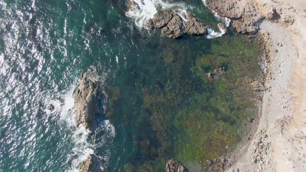 Top-down coastal water with rock outcrops, foamy waves, and sandy shore.