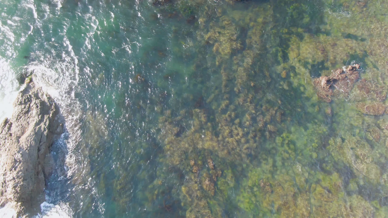 A central rocky outcrop in Bahia Ascuncion sits in clear green-blue water as white foam curls around its edges, with no visible people.
