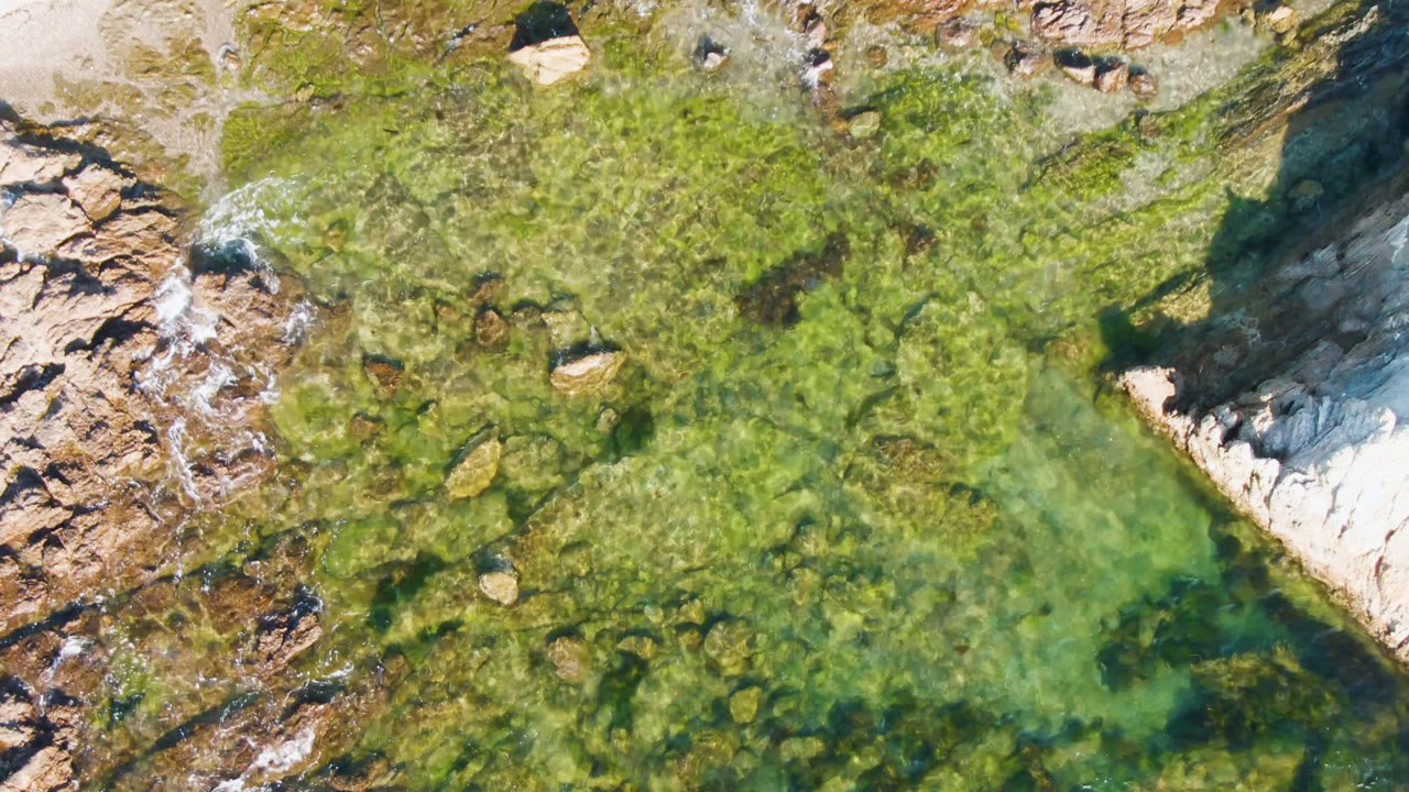 At 0:01, the overhead view in Bahia Ascuncion centers more of the rocky shelf, with a small dark hole near pale sand and no visible people.