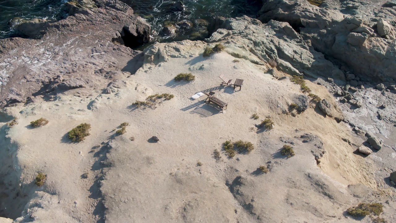 Overhead frame from Bahia Ascuncion, Baja California Sur, Mexico, showing two empty chairs on pale sand below rocky shoreline and dark water, with no visible people.