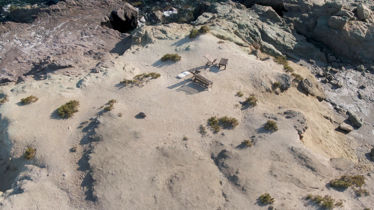 Aerial frame from Bahia Ascuncion, Baja California Sur, Mexico, with two empty chairs casting long shadows on sand beside scattered shrubs and shoreline rocks, with no visible people.