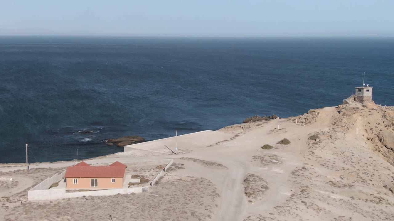 Aerial view of a sandy coastal hill with a red-roofed building and ocean beyond.
