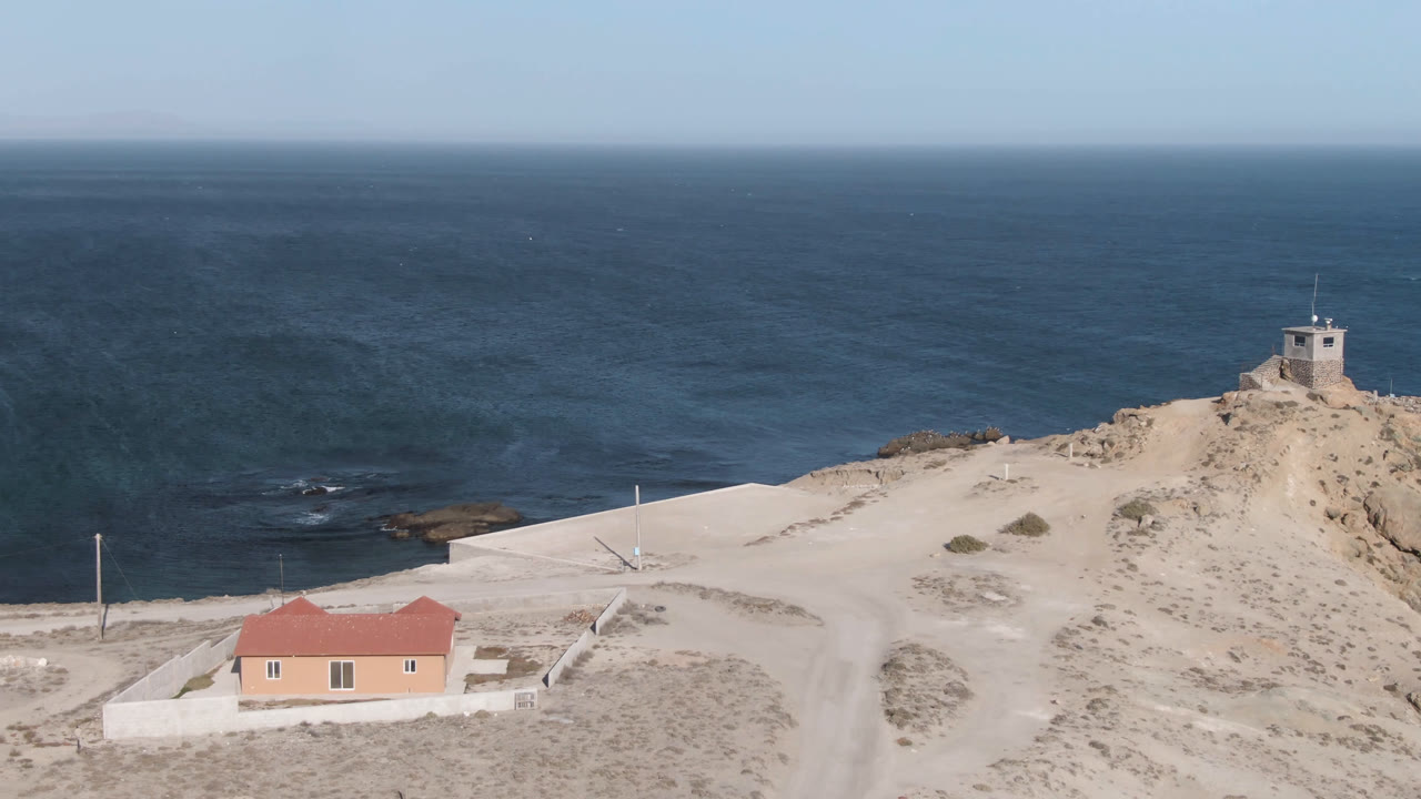 One second into the Bahia Ascuncion clip, the sandy slope and road sit below a small hilltop structure, with the red-roofed building partly lower left and no visible people.
