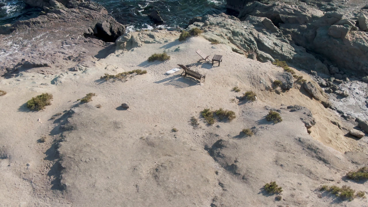 Elevated view in Bahia Ascuncion, Baja California Sur of a wooden bench and chair on pale sand above rocky surf, with no visible people.