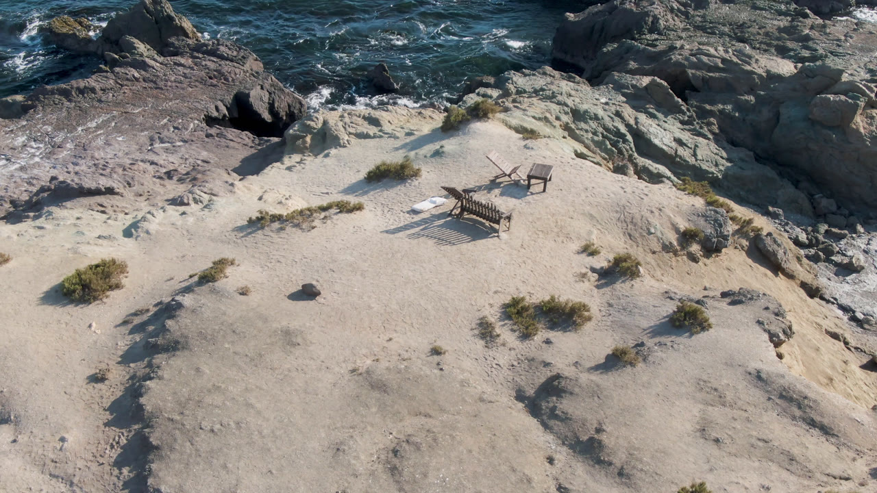 Aerial-looking frame of the Bahia Ascuncion beach bench and chair, with more foamy water visible along the rocks and no visible people.