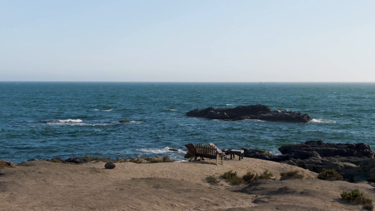 At 0:00 in Bahia Ascuncion, Baja California Sur, a sandy foreground with a wooden bench faces blue ocean water, dark rocks, and a pale sky with no visible people.