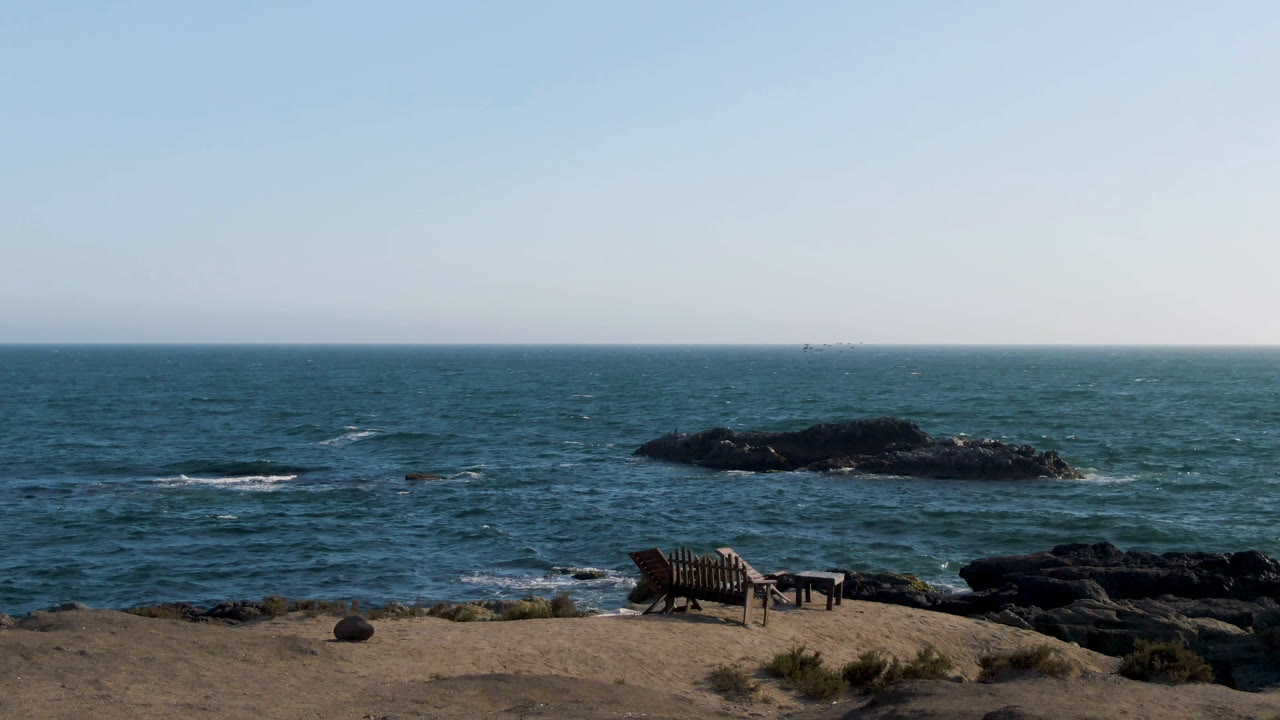 At 0:01 in Bahia Ascuncion, Baja California Sur, the bench and small table sit near the lower edge while dark shoreline rocks and low waves stretch across the middle distance with no visible people.