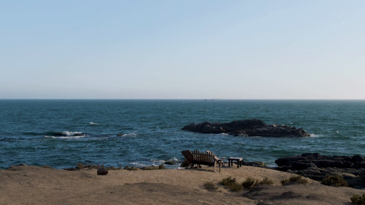 Wide ocean view with a bench on sand and black rocks offshore.