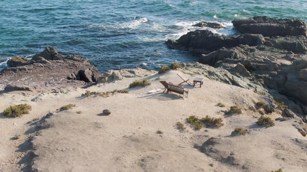 Elevated view of Bahia Ascuncion beach with two chairs and a small table on pale sand, dark rocks near the surf, blue ocean beyond, and no visible people.