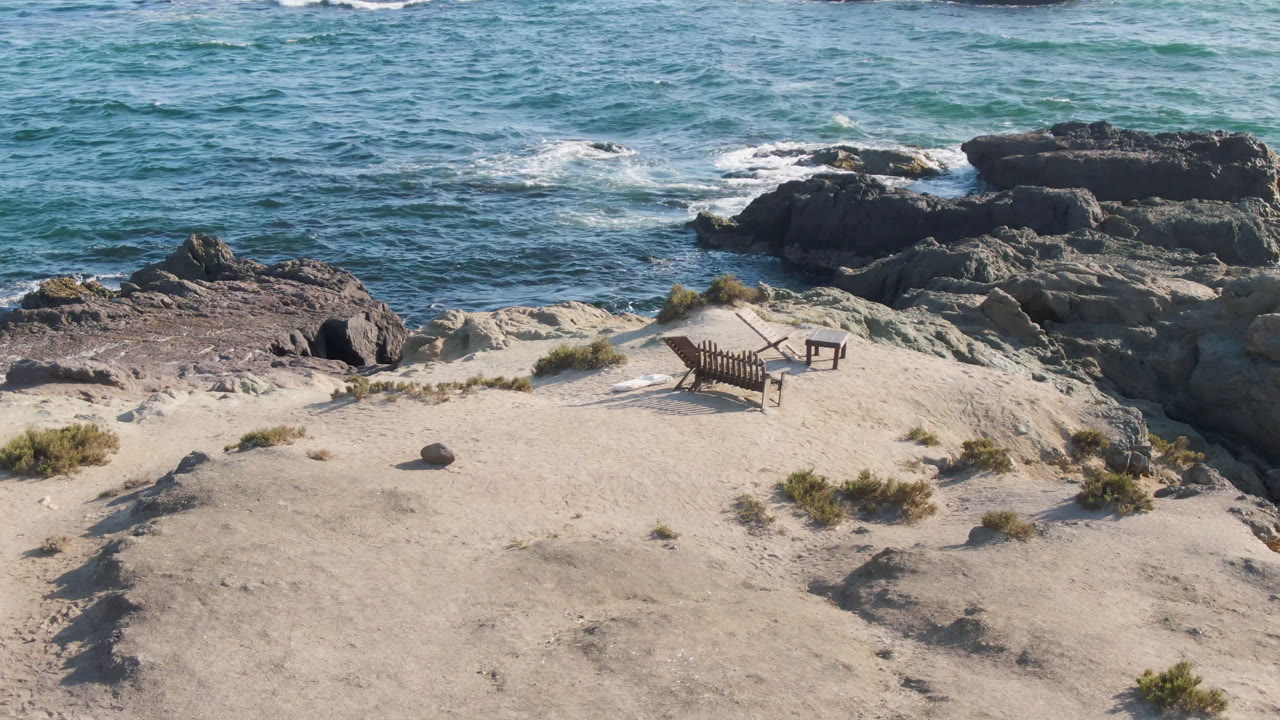 A slightly later elevated frame shows the chairs and table centered on the sandy rise at Bahia Ascuncion, with rocky shoreline and foam lines in the water behind them, no visible people.