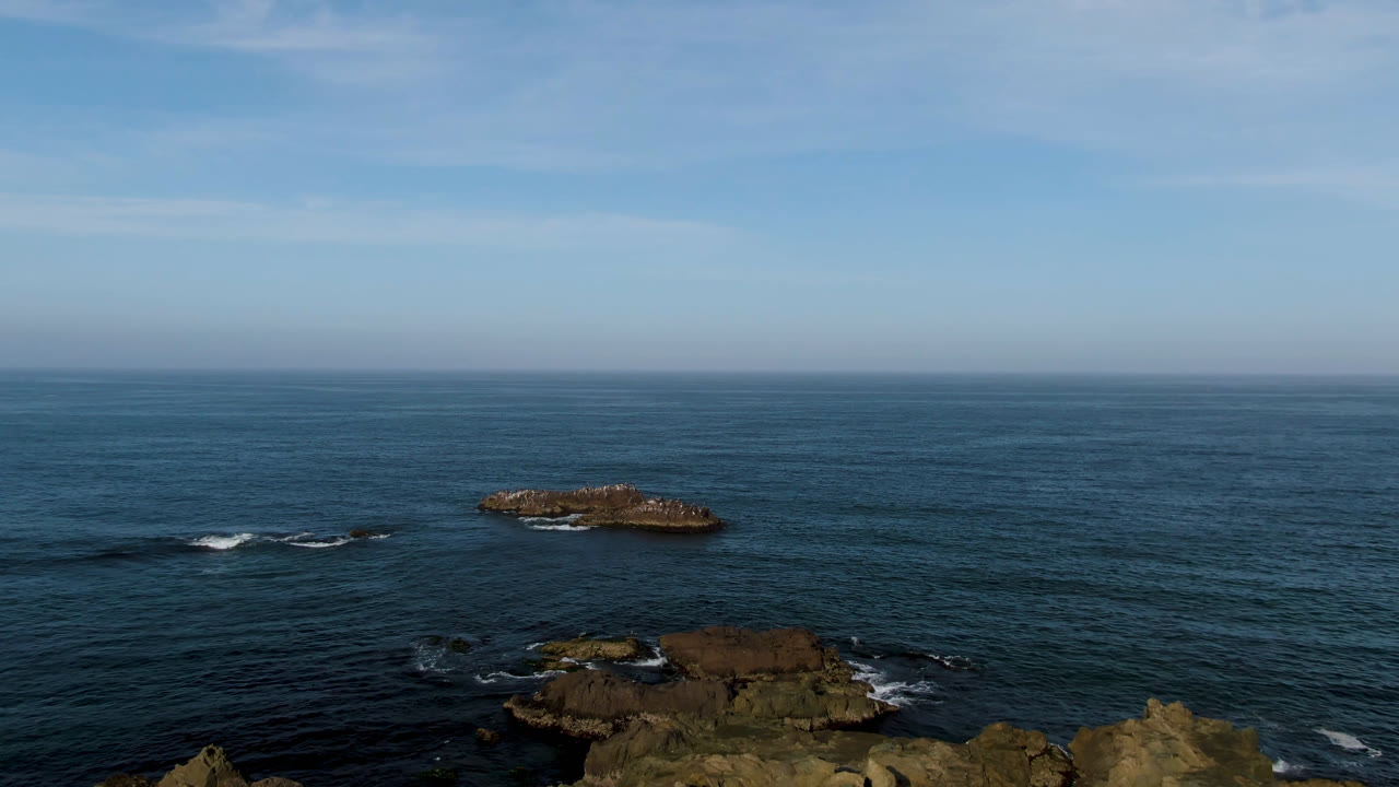 Wide aerial view in Bahia Ascuncion, Baja California Sur, Mexico, showing open blue ocean, two offshore rock formations, rocky shoreline at the bottom edge, and no visible people.
