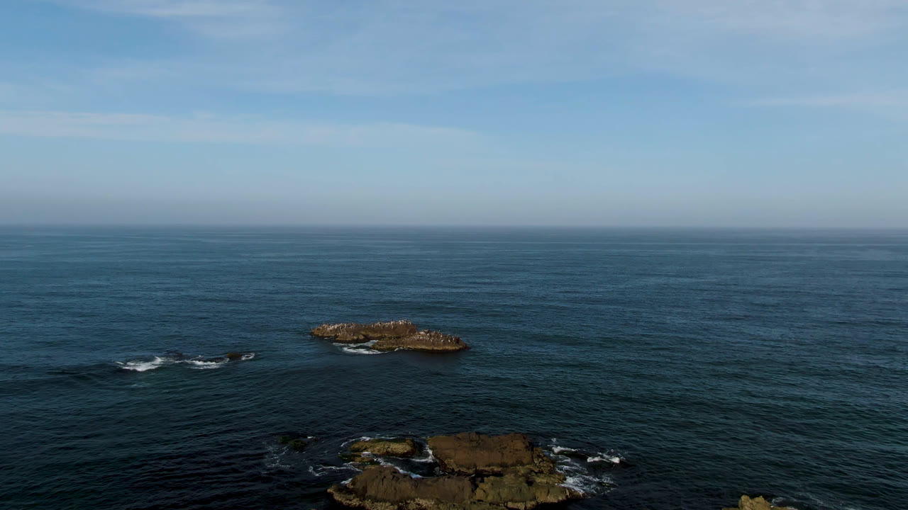 Aerial frame from Bahia Ascuncion with the offshore rocks slightly shifted across the wide ocean composition, small white wave breaks near the lower rocks, and no visible people.