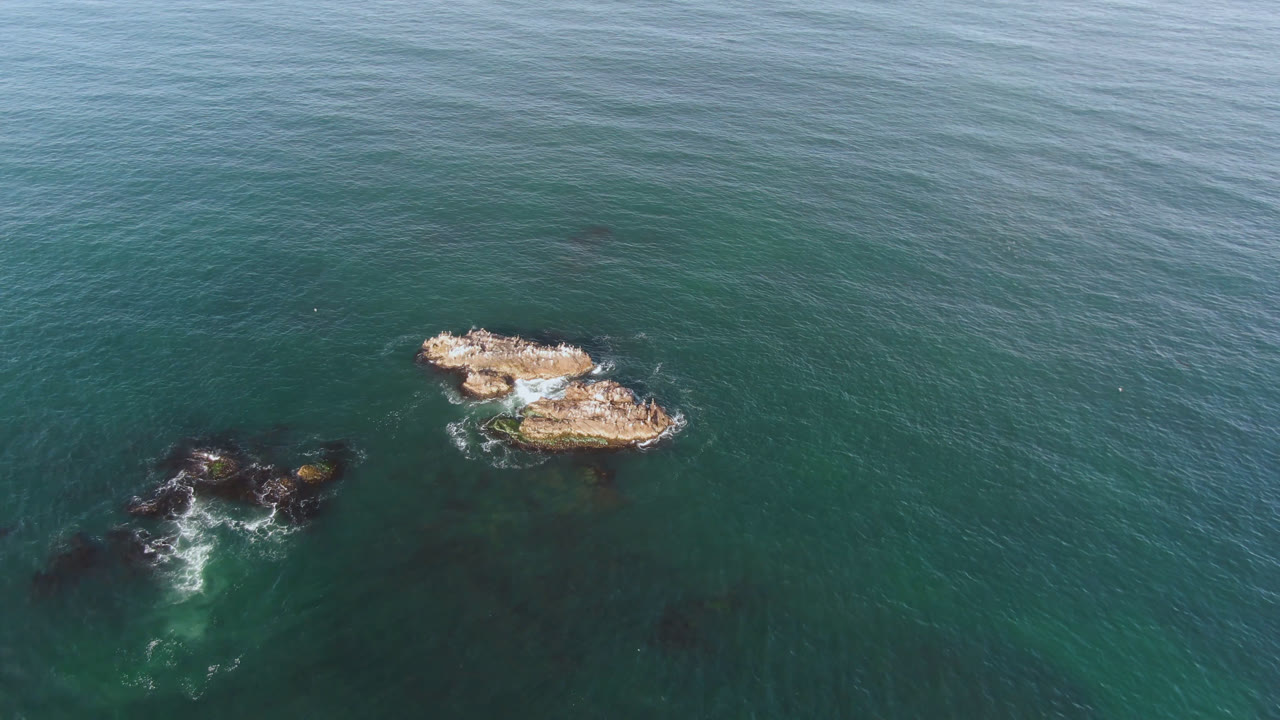 Overhead frame from Bahia Ascuncion, Baja California Sur, Mexico, showing two tan rock clusters in green-blue water with white surf and no visible people.