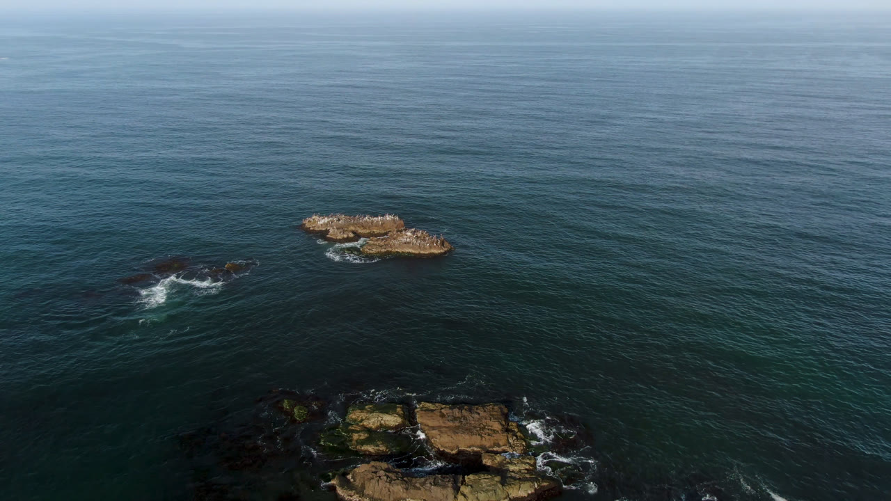 High aerial frame over Bahia Ascuncion, Mexico, showing two offshore rock formations in teal water, shoreline rocks at the bottom edge, and no visible people.