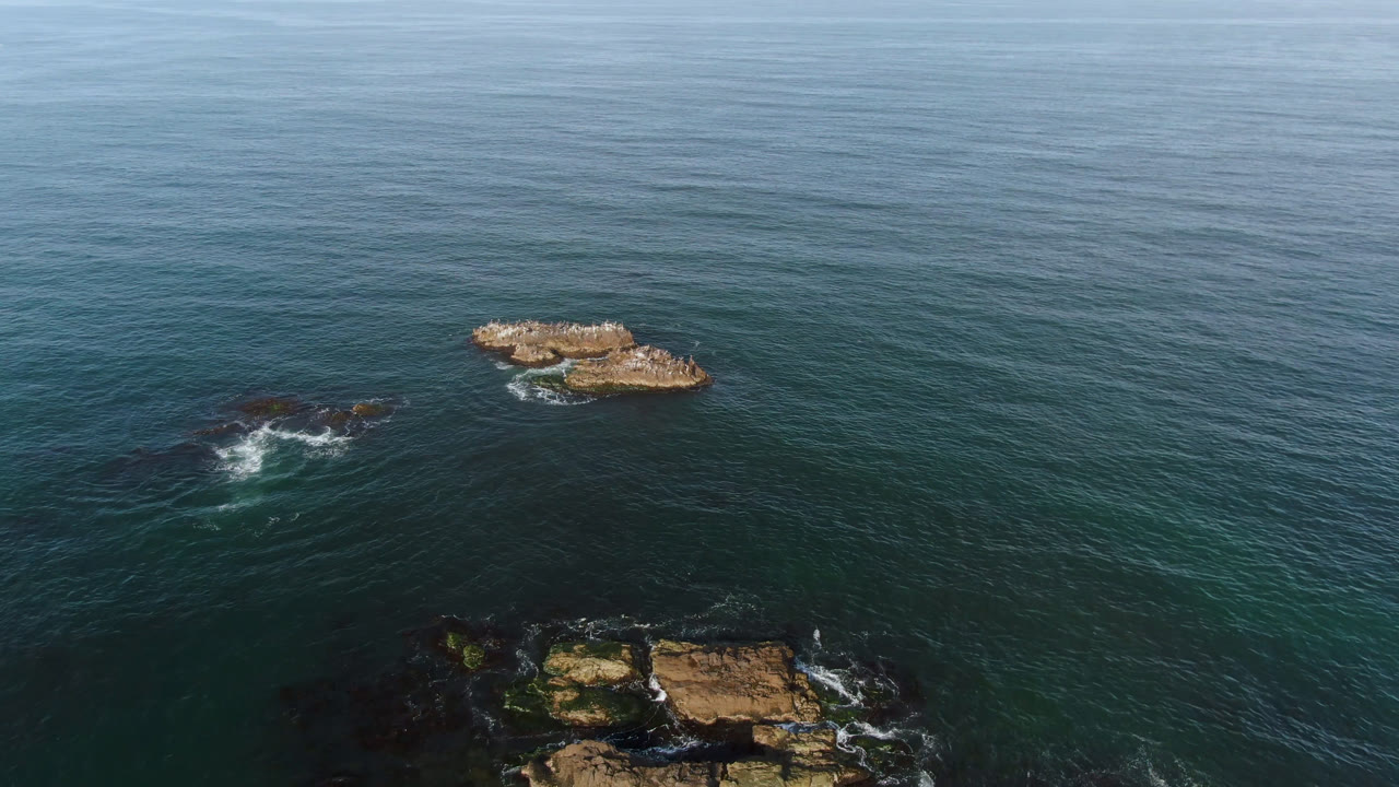 Aerial view in Bahia Ascuncion, Mexico, with the larger tan rock to the right, a darker rock patch to the left, open water across the background, and no visible people.