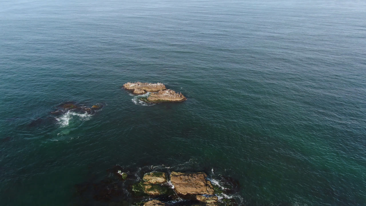 Drone view of rocky outcrops with surf in green-blue water.