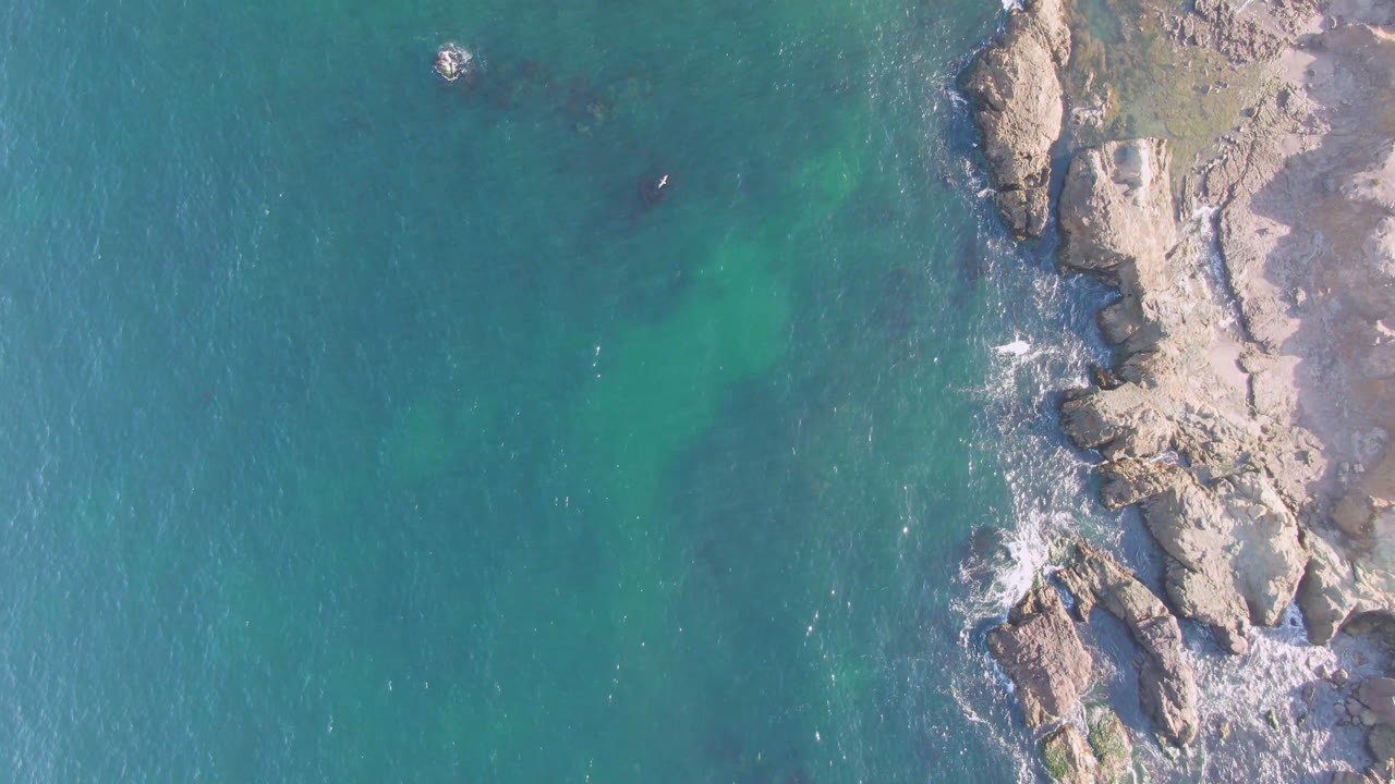 Top-down aerial frame over Bahia Ascuncion, Baja California Sur, with turquoise water on the left, pale rock shelves on the right, small surf marks, and no visible people.