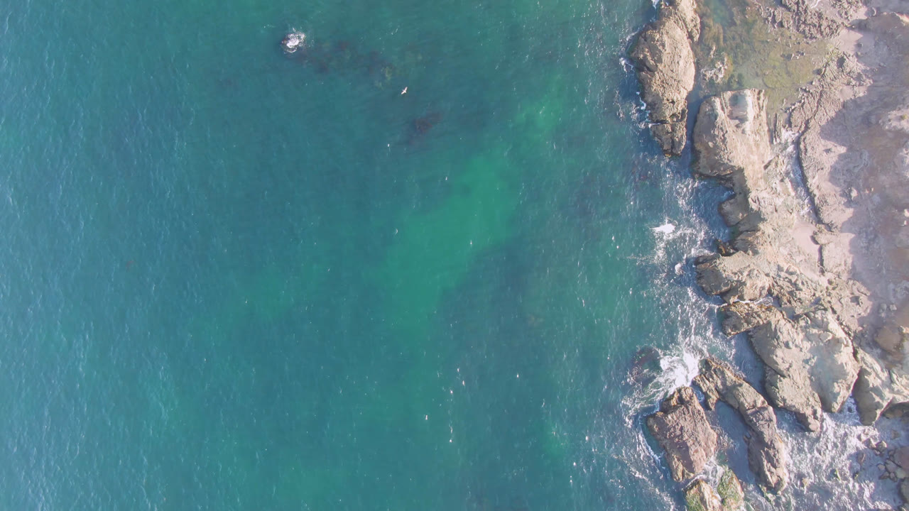 Aerial view over Bahia Ascuncion with blue-green water dominating the left side, a vertical rocky outcrop on the right edge, dark underwater patches, and no visible people.