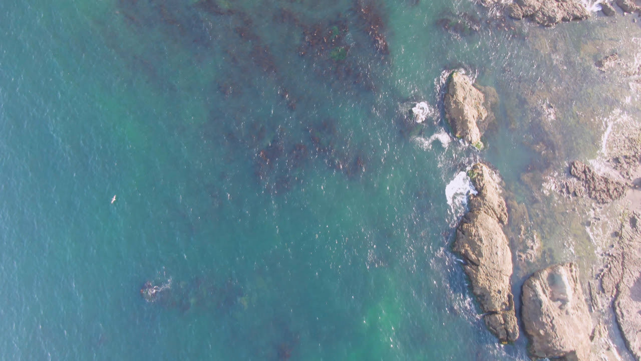 Overhead view of turquoise water and tan shoreline rocks at Bahia Ascuncion, with small foam breaks around the stones and no visible people.