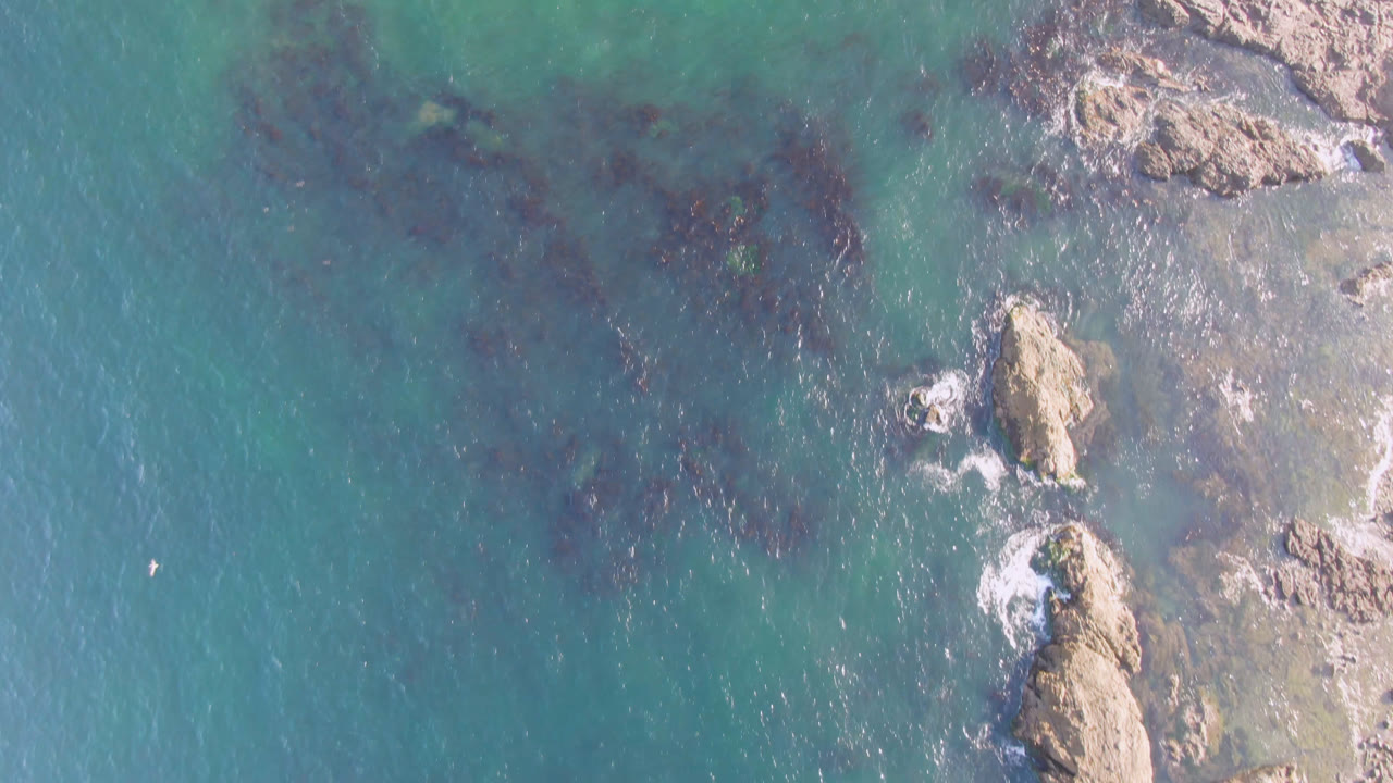 Aerial frame from Bahia Ascuncion showing green-blue water, dark underwater patches, right-edge rocks, and no visible people.