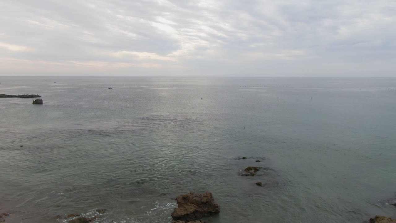 Aerial view over Bahia Ascuncion, Baja California Sur, Mexico, with a small boat near the left waterline, rocks in the lower frame, a pale cloudy horizon, and no visible people.