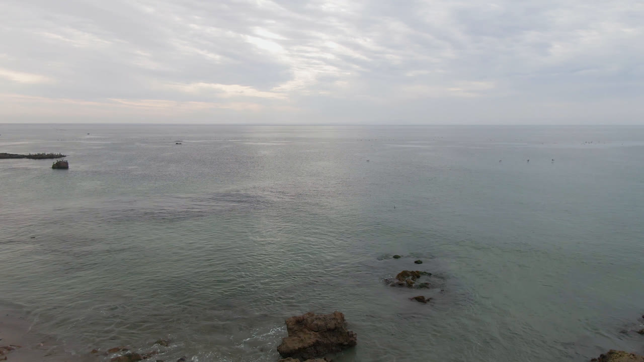 Wide aerial frame in Bahia Ascuncion showing the small boat left of center, scattered rocks near the lower edge, calm gray-green water, a distant horizon, and no visible people.