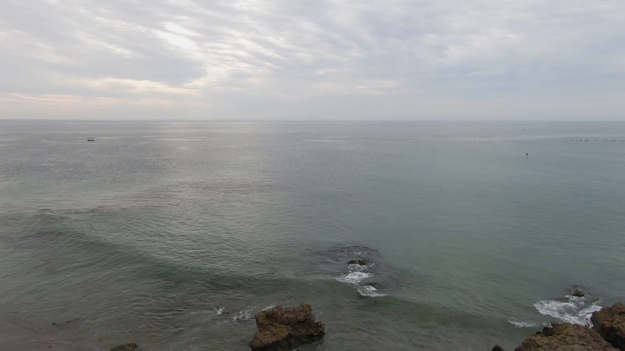 Bahia Ascuncion ocean surface stretches to a pale horizon while foreground rocks sit near the lower edge and faint white surf breaks nearby with no visible people.