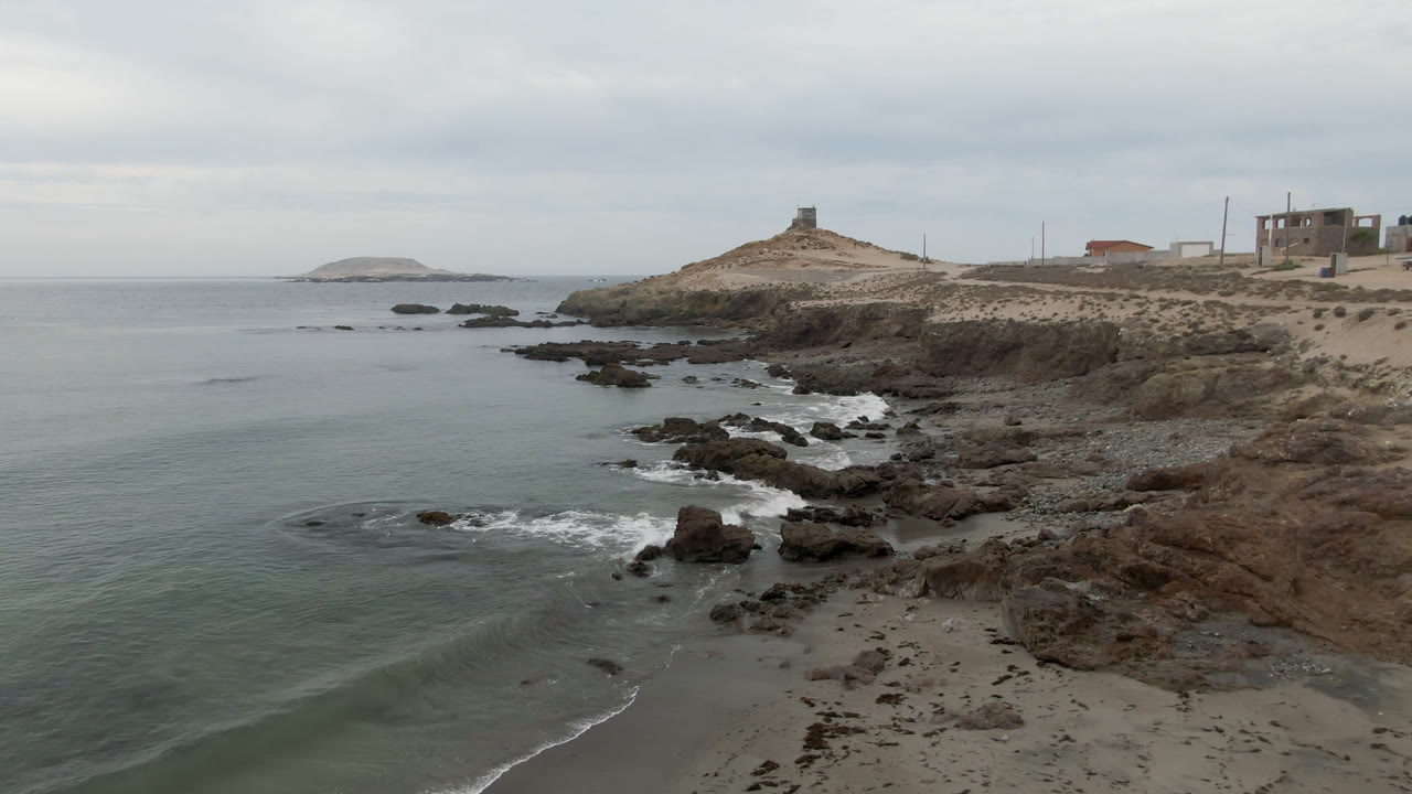 Rocky shoreline with ocean, distant lighthouse, and cloudy sky.
