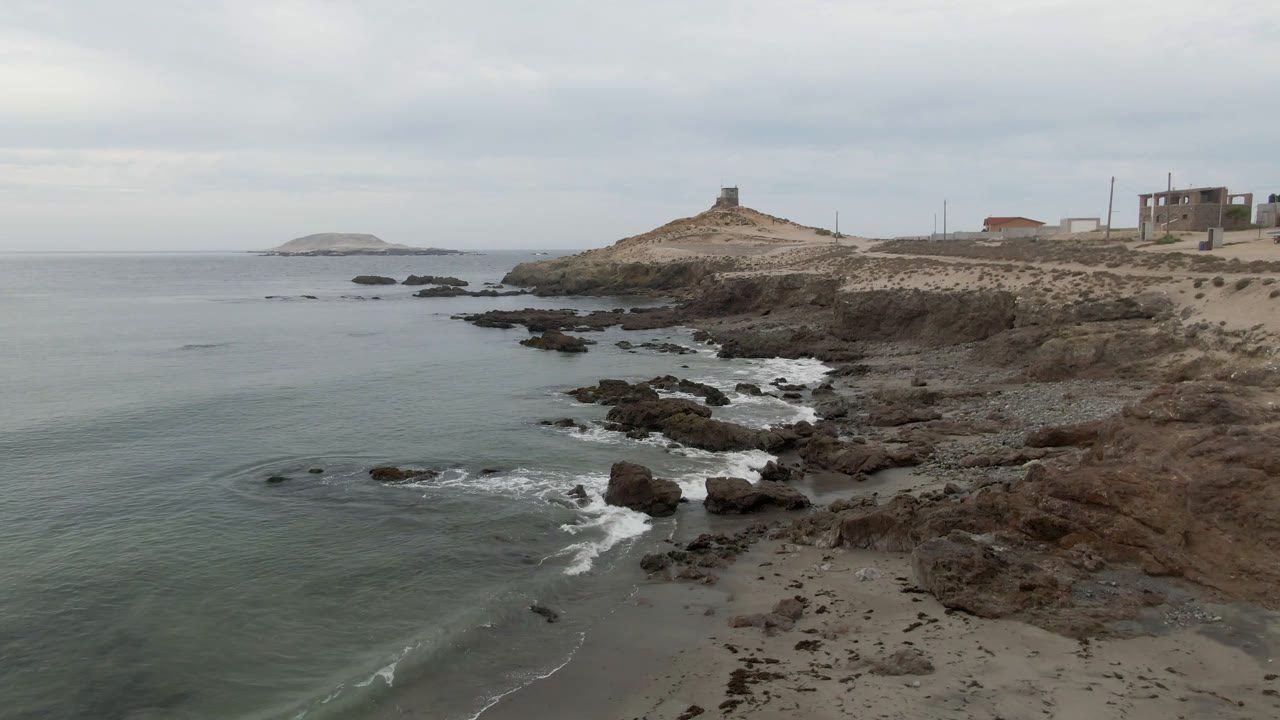 Elevated coastal frame in Bahia Ascuncion showing green-gray water, scattered dark rocks, white surf lines, the lighthouse on the far headland, and no visible people.