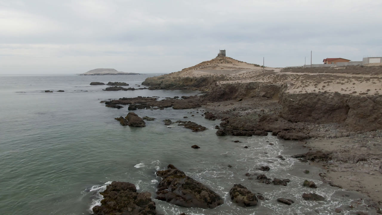 Bahia Ascuncion coastline with shallow water around scattered rocks, pale beach below the headland, the lighthouse near the horizon, and no visible people.
