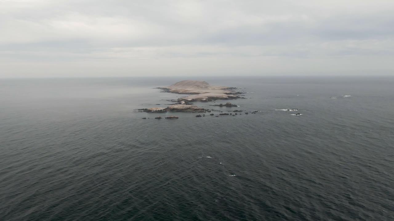 Aerial view in Bahia Ascuncion, Baja California Sur, with a rocky island centered beyond dark rippled ocean, white wave breaks near the rocks, a hazy horizon, and no visible people.