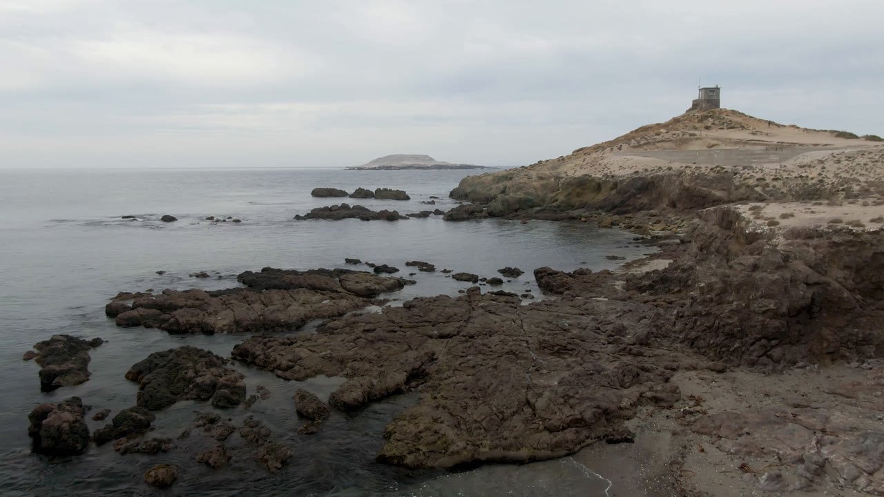 Elevated view of Bahia Ascuncion rocks and shallow ocean water with a small lighthouse on the sandy hill at right and no visible people.