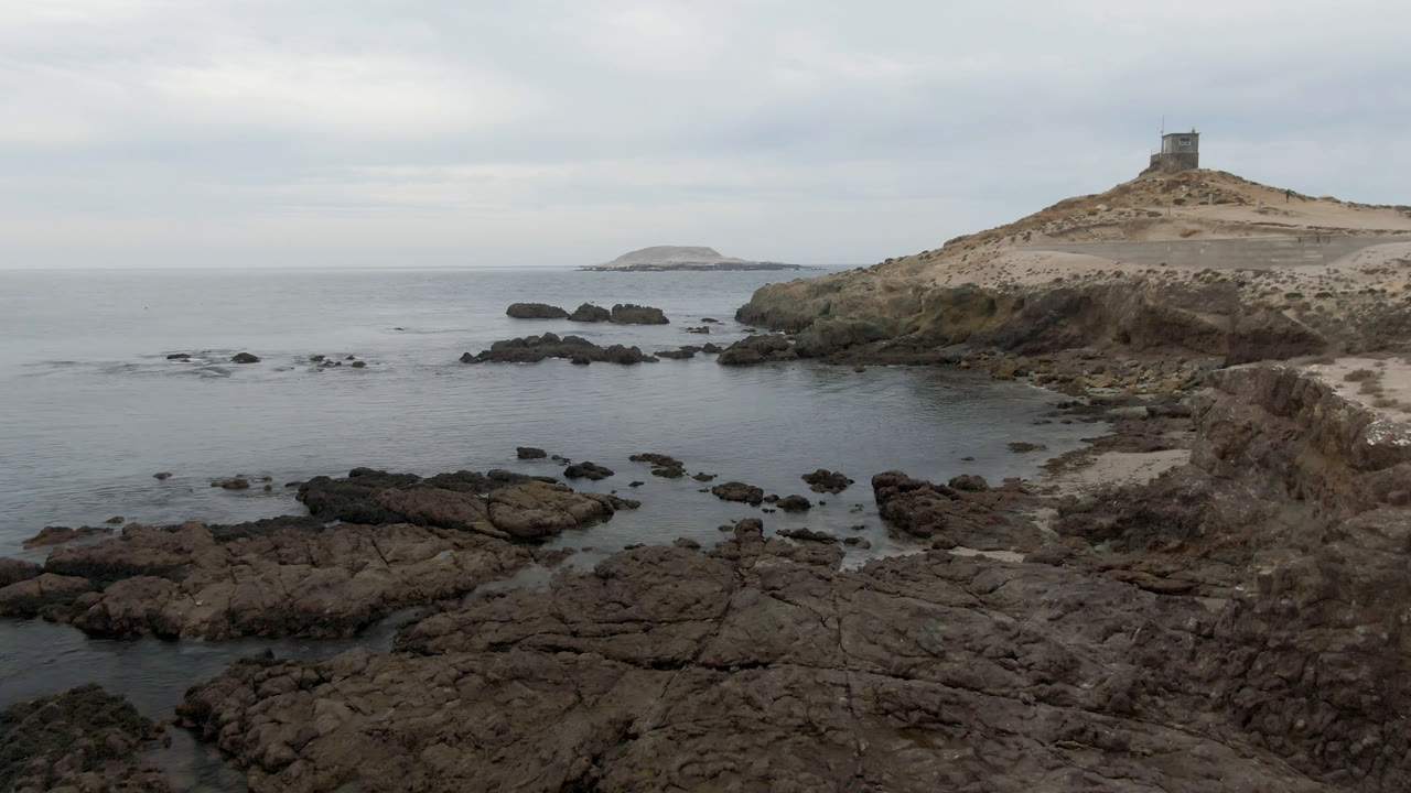 Bahia Ascuncion ocean water spreads around dark rock outcrops with the lighthouse hill on the right and no visible people.