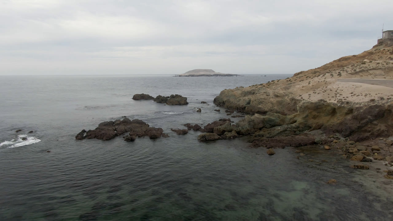 Elevated view of Bahia Ascuncion coastline with dark rocks in the foreground, a scrub-covered headland at right, an offshore island on the horizon, and no visible people.
