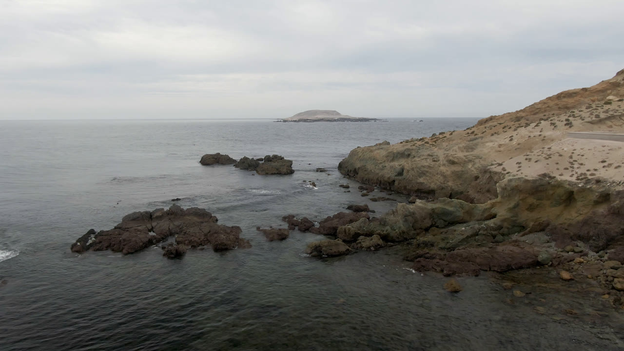 Bahia Ascuncion coastal rocks sit in dark water beside a tan headland, with a low offshore island centered beyond the horizon and no visible people.