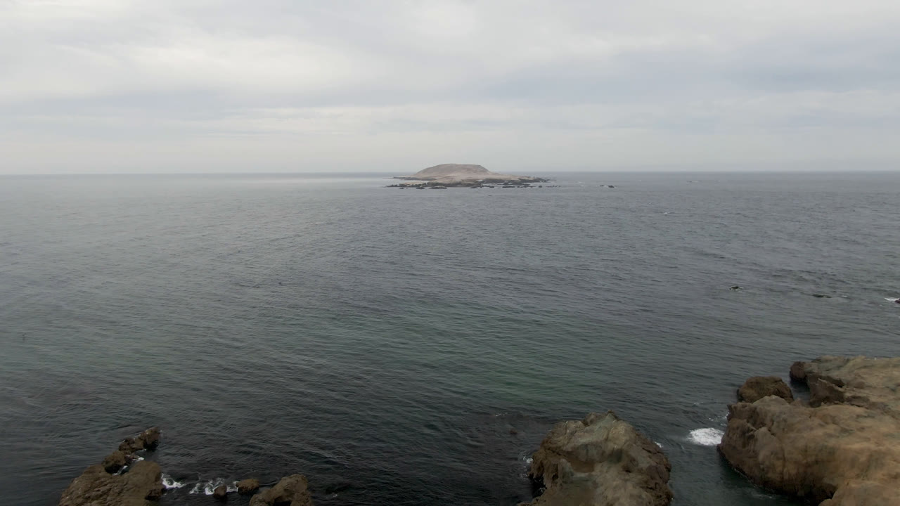 Bahia Ascuncion coastline seen from above with larger foreground rock outcrops, textured grey-blue ocean, the distant island centered near the horizon, and no visible people.