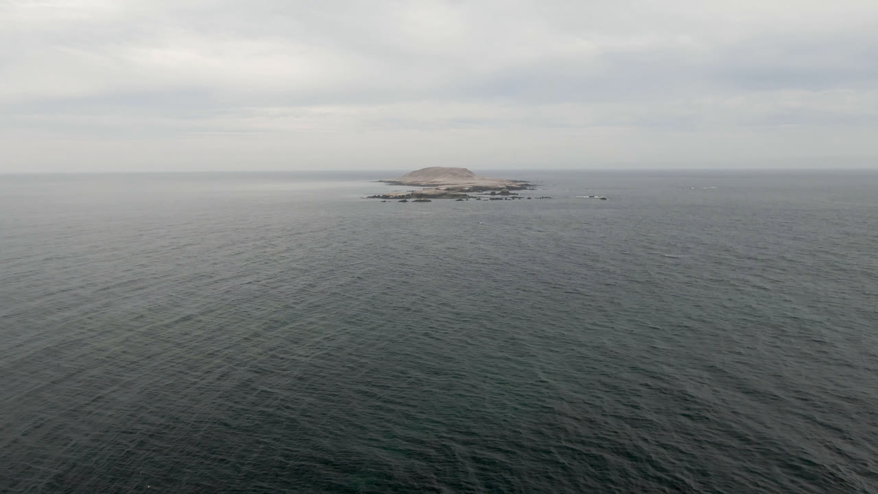 Bahia Ascuncion aerial frame with rippled ocean across most of the view, the small island held slightly left of center near the horizon, low rocks around it, and no visible people.