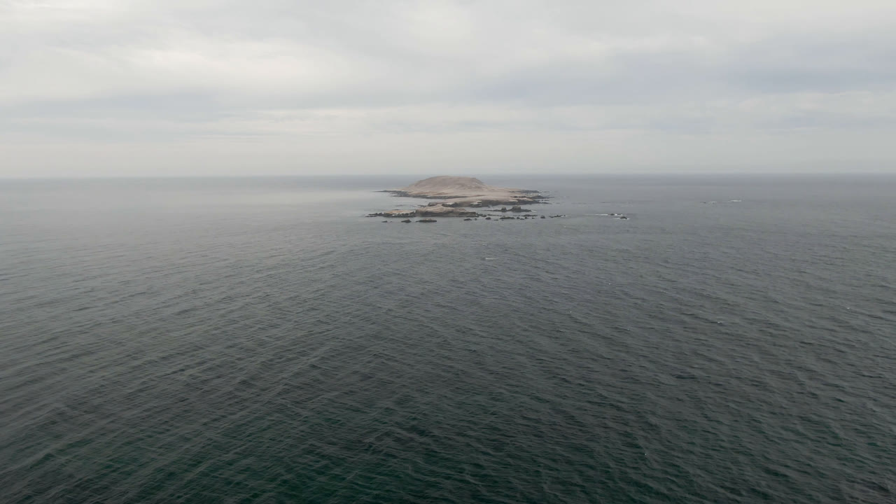Opening frame of Bahia Ascuncion ocean with a distant low rocky island, gray cloud layers, rippled dark water in the foreground, and no visible people.